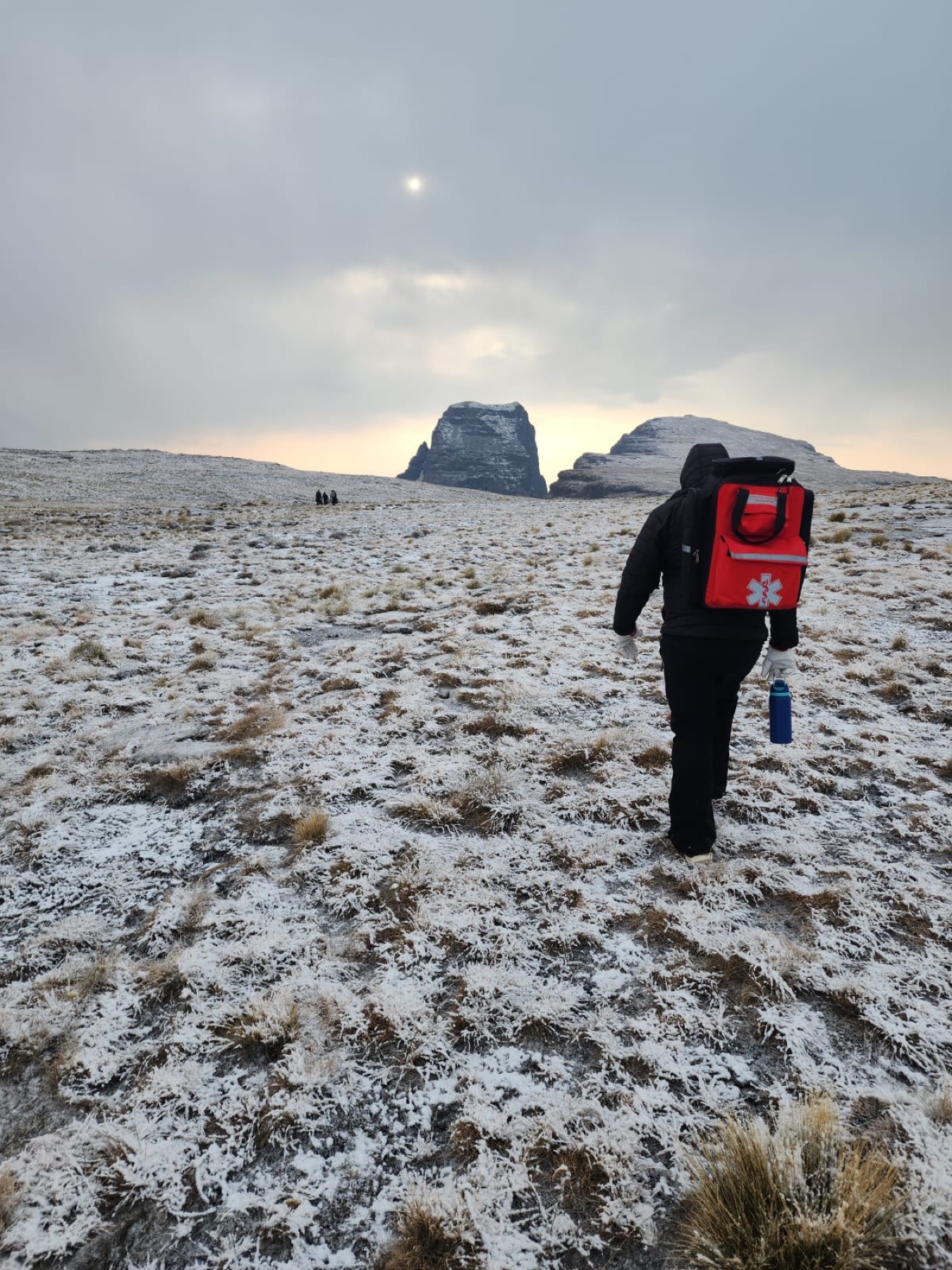 Medic carrying a red medical backpack across a snow-covered mountain landscape